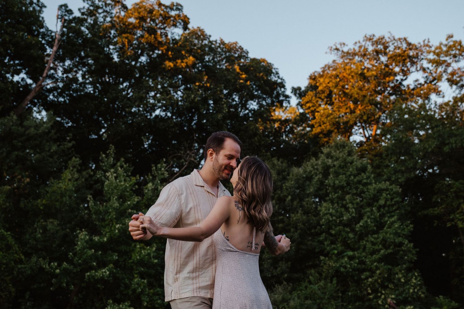 Happy couple dancing outdoors at sunset surrounded by lush green and golden autumn trees.