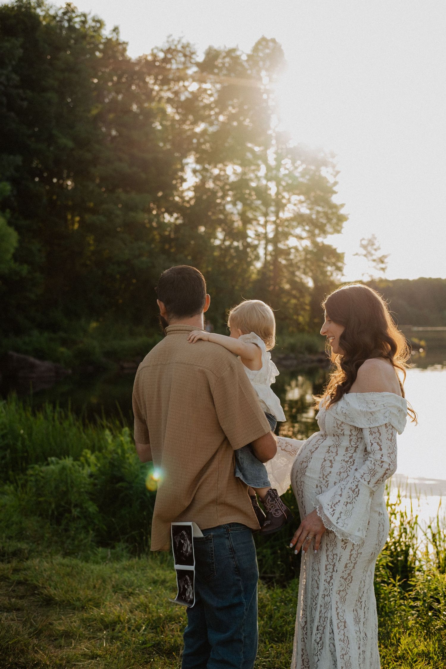 Pregnant woman in white lace dress with partner holding toddler, standing by a sunlit lake at golden hour.