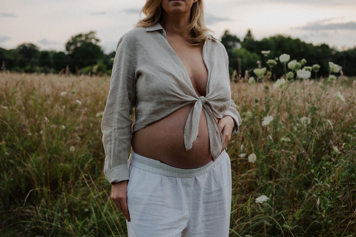 Pregnant woman in tied linen shirt and white pants standing in a wildflower field, showcasing her baby bump.