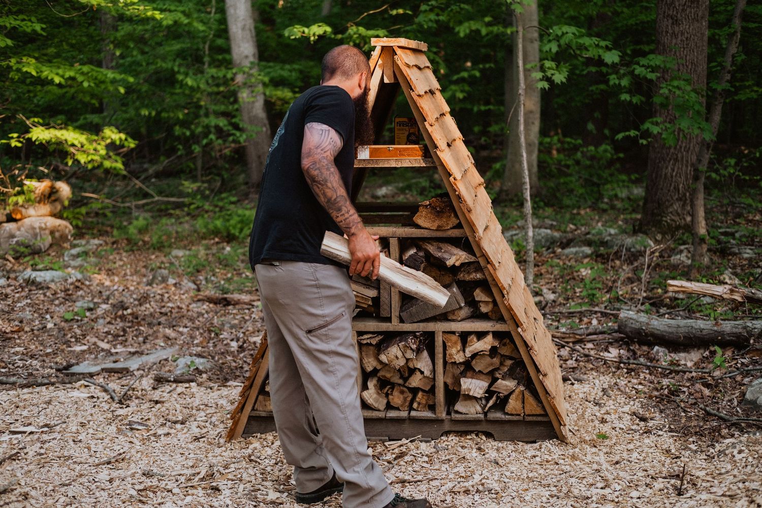 Man stacking firewood in a triangular A-frame outdoor wood storage structure in a forest setting.