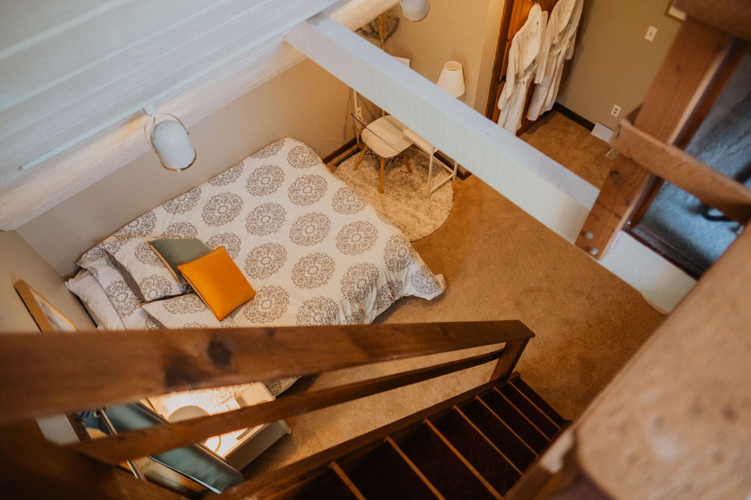 Aerial view of a cozy loft bedroom with patterned bedding, orange accent pillow, and wooden staircase railing.
