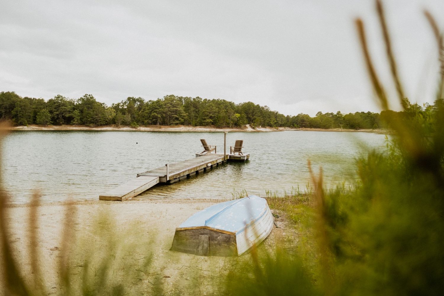 A wooden dock with Adirondack chairs overlooks a calm lake, with a rowboat resting on a grassy shore nearby.