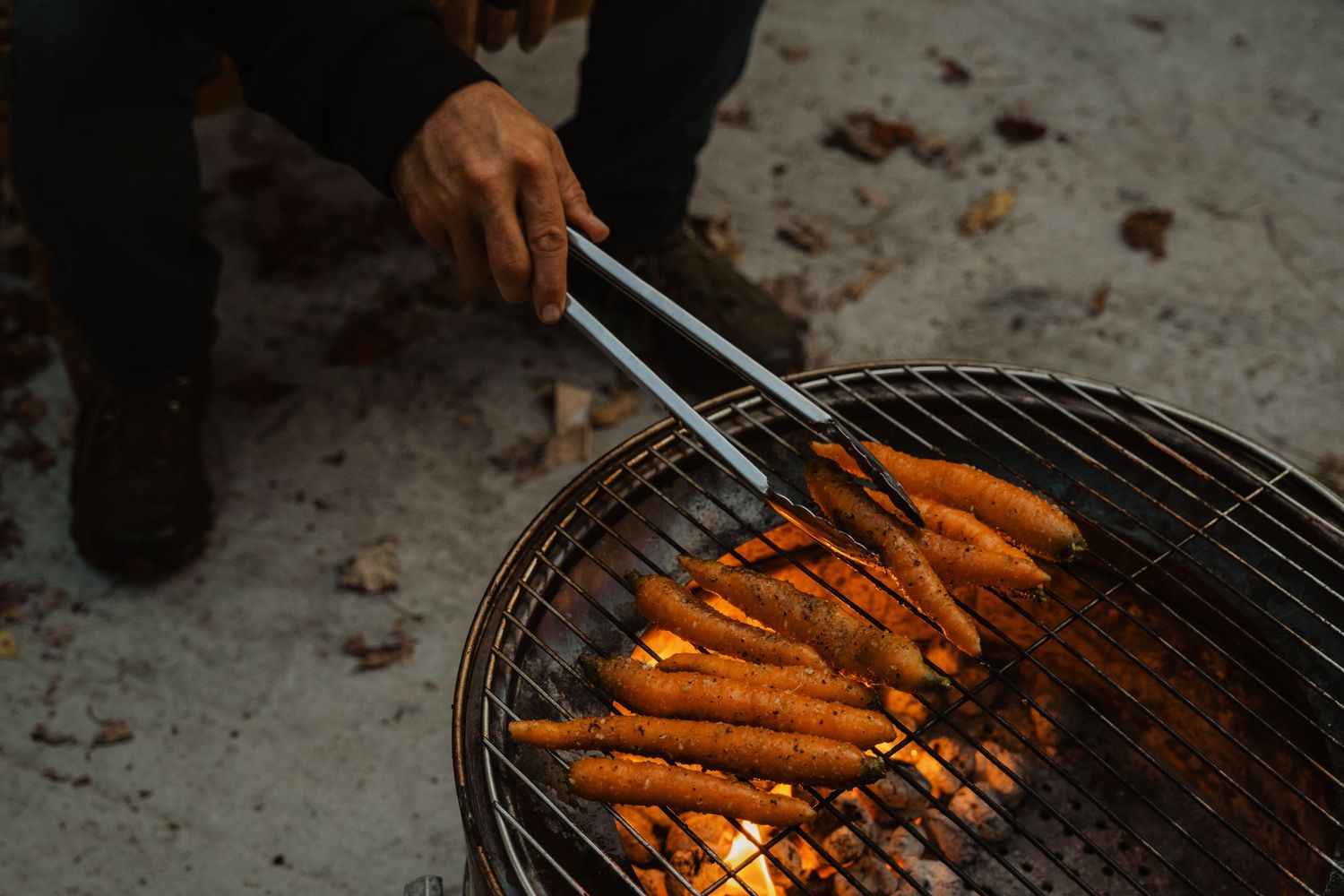 Person grilling fresh carrots on a round charcoal grill using metal tongs over open flames outdoors.