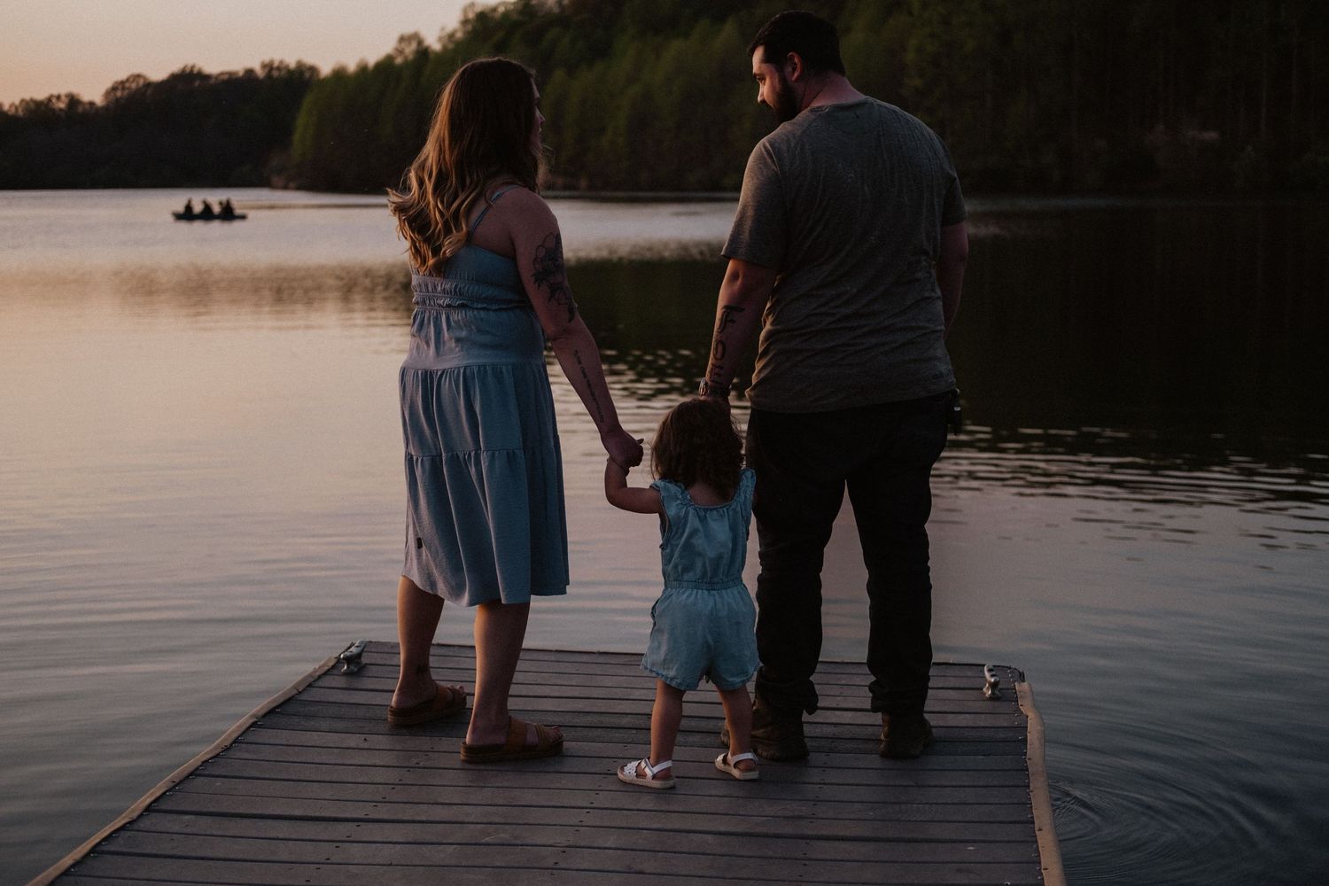 Family of three holding hands on a wooden dock at sunset overlooking a calm lake.