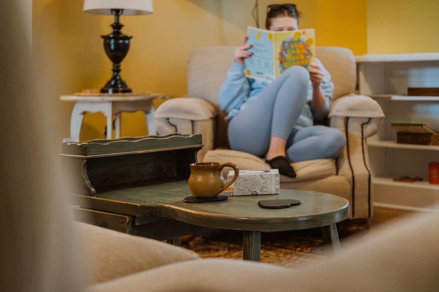 Woman relaxing on armchair reading a colorful book in a cozy vintage living room with yellow walls.
