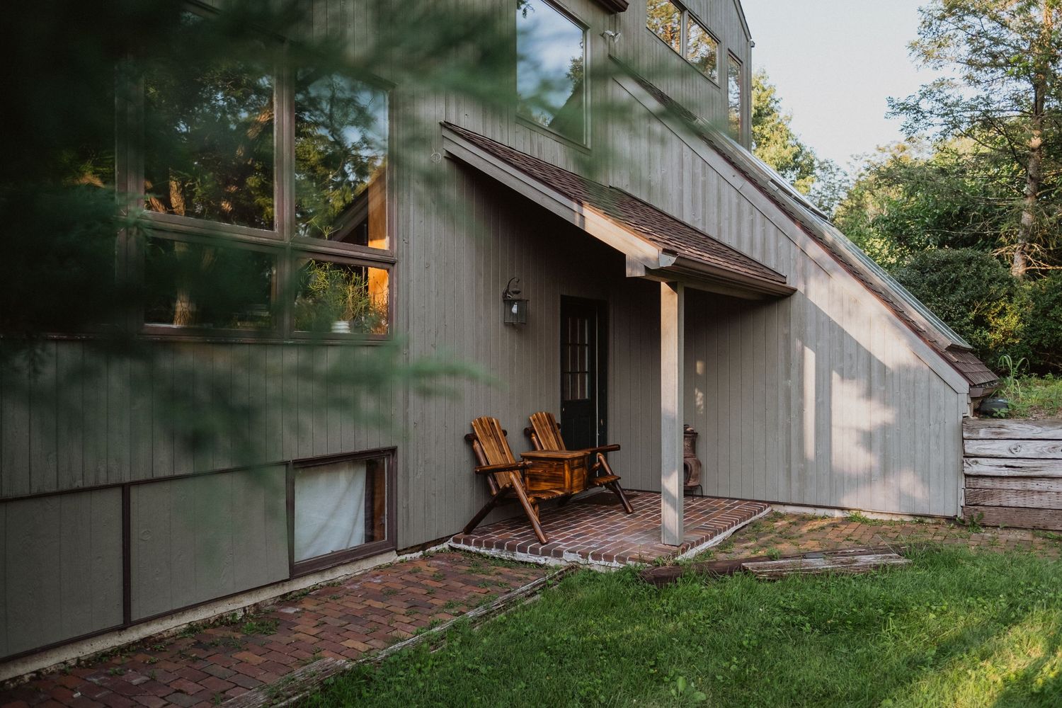 Modern wood-sided home with Adirondack chairs on brick patio surrounded by lush green trees.