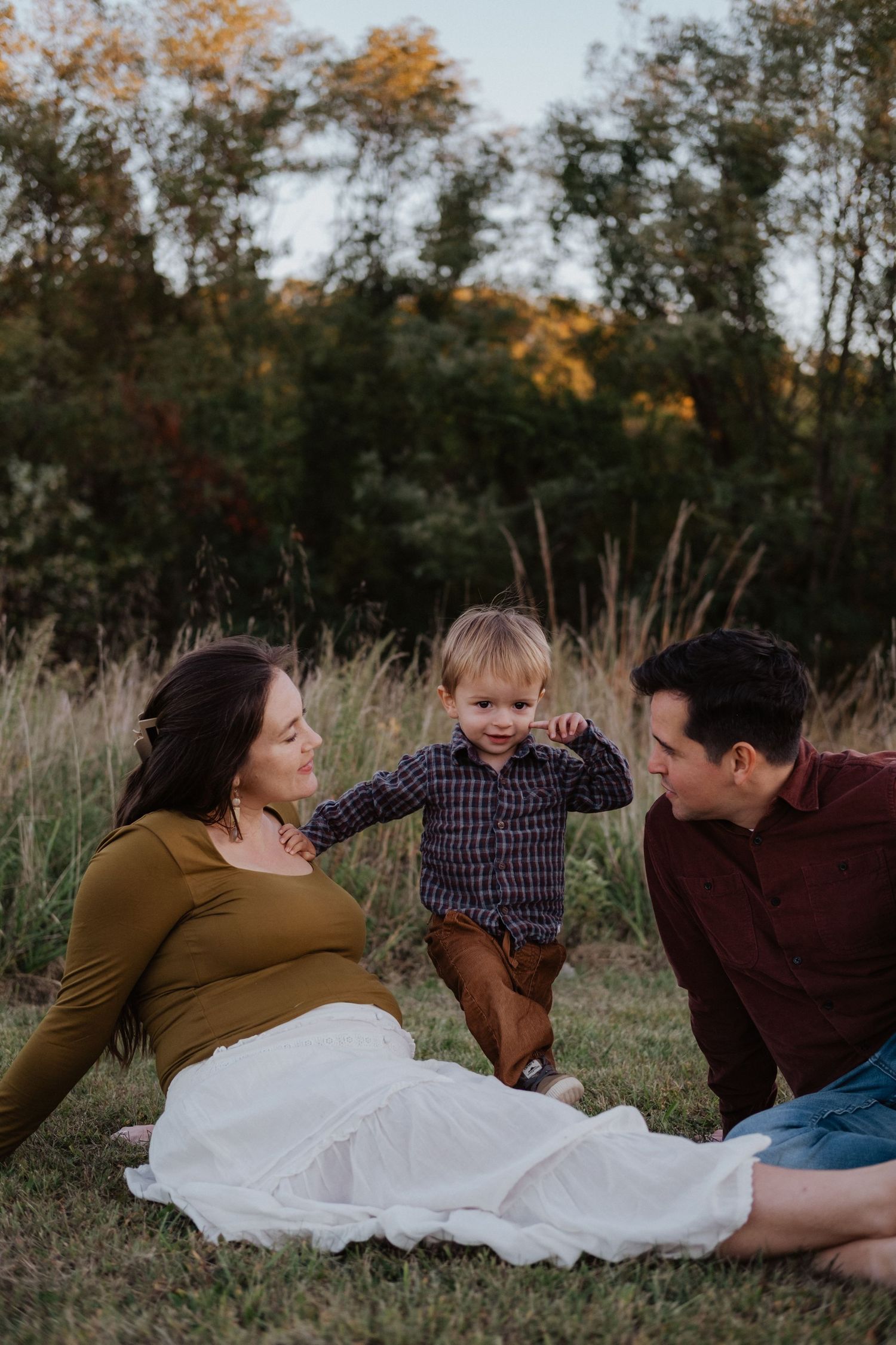 Pregnant mother relaxing on grass with toddler son and partner during fall maternity session.
