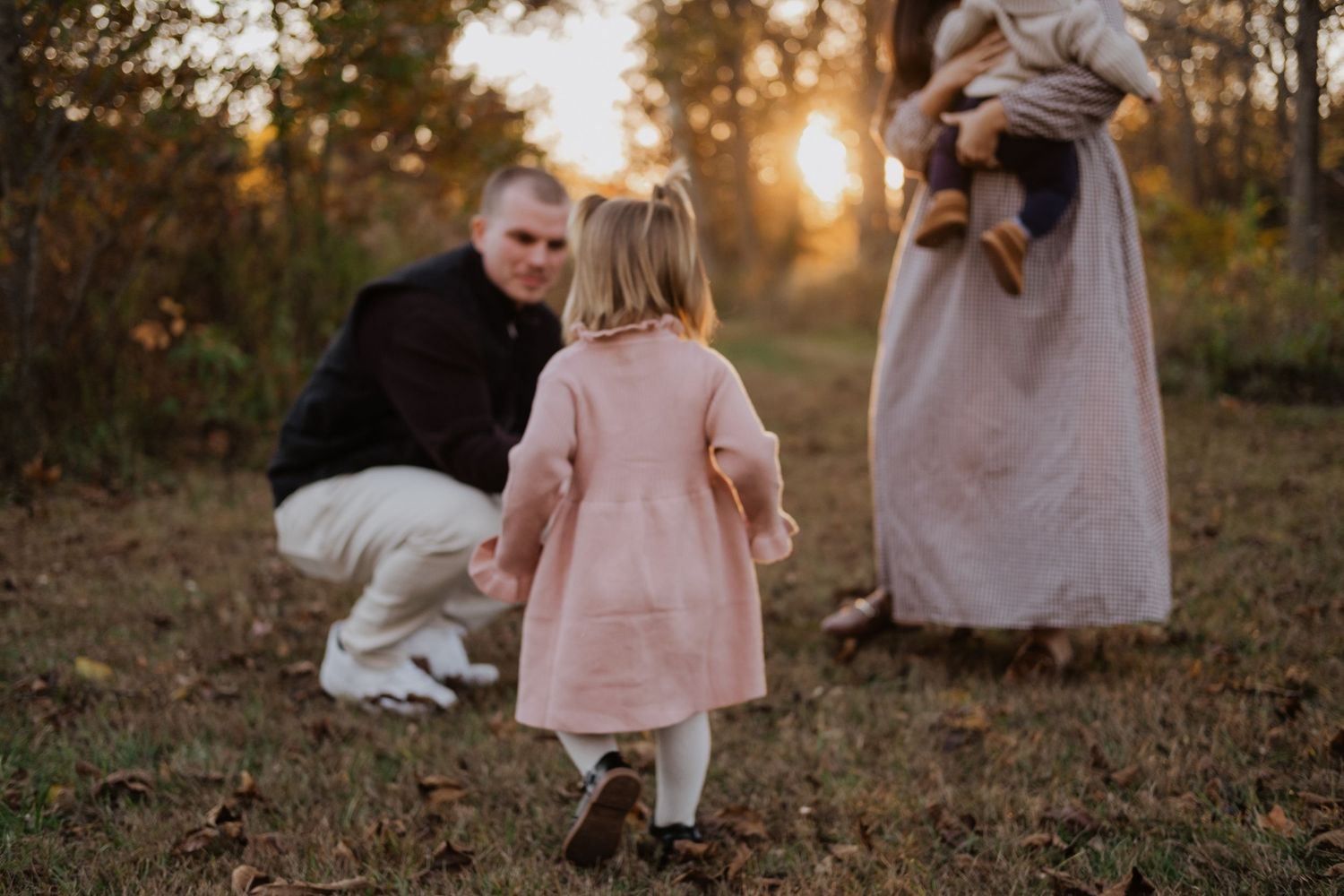 Little girl in pink coat walking toward father crouching at sunset in autumn woods.