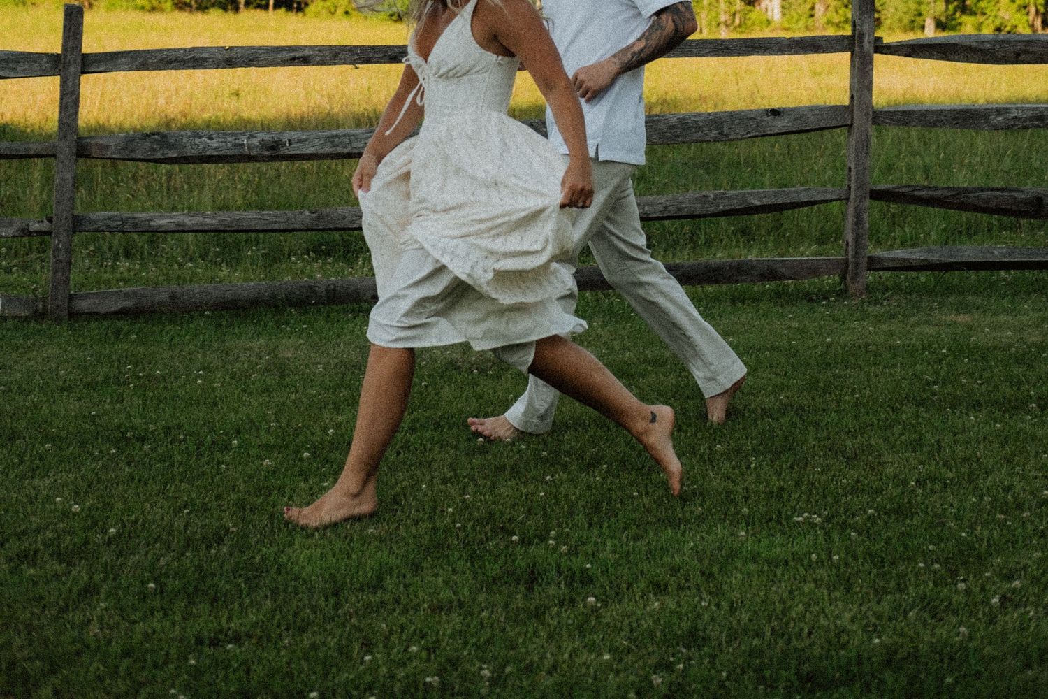 Barefoot couple running together on green grass near a rustic wooden fence in a romantic outdoor setting.