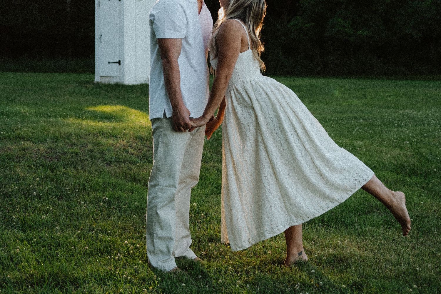 Couple holding hands in a green field, woman in flowing white dress lifts foot playfully near a white barn.
