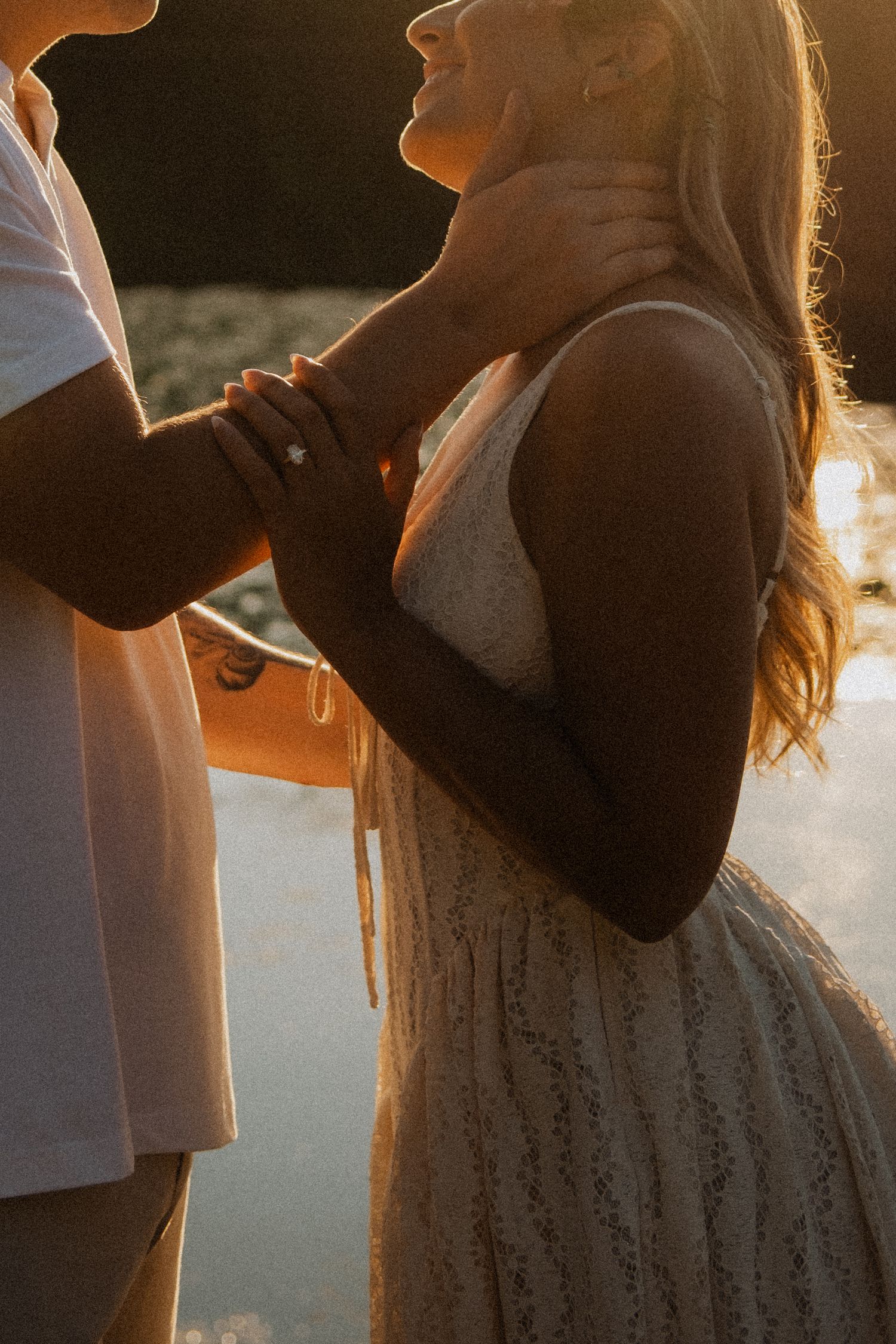 Couple sharing a romantic sunset moment, woman in white lace dress with engagement ring, bathed in golden light.