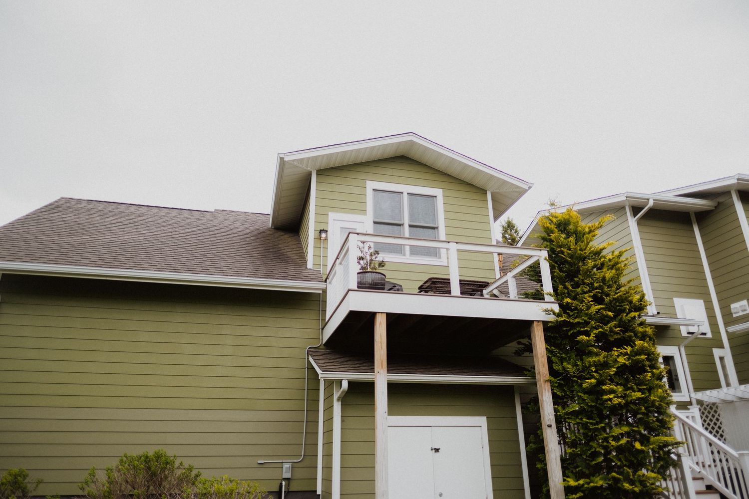 Green two-story house with white balcony, brown shingle roof, and white garage door on overcast day.