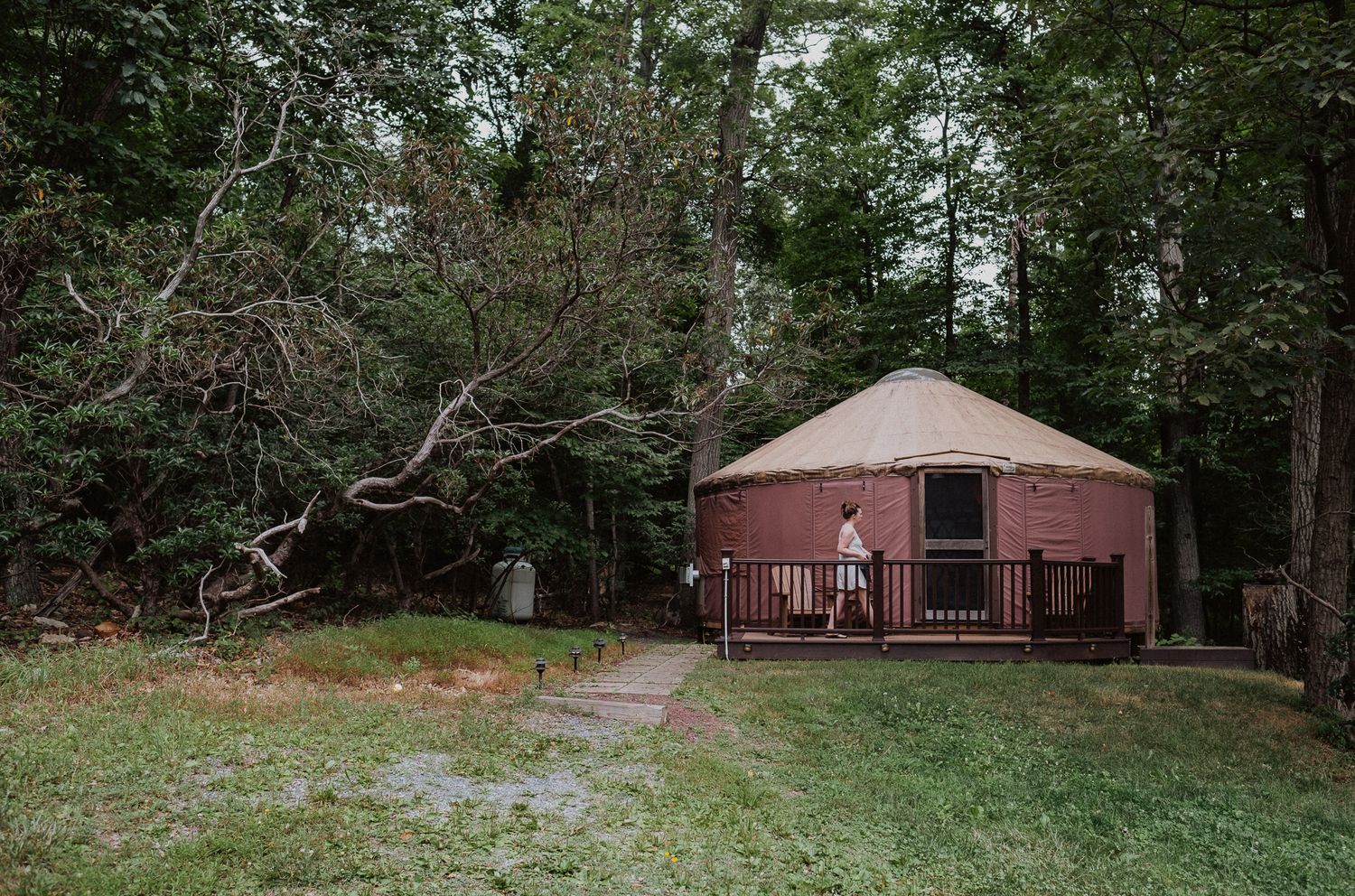 A person stands on the deck of a rustic red yurt surrounded by tall trees in a wooded campsite setting.