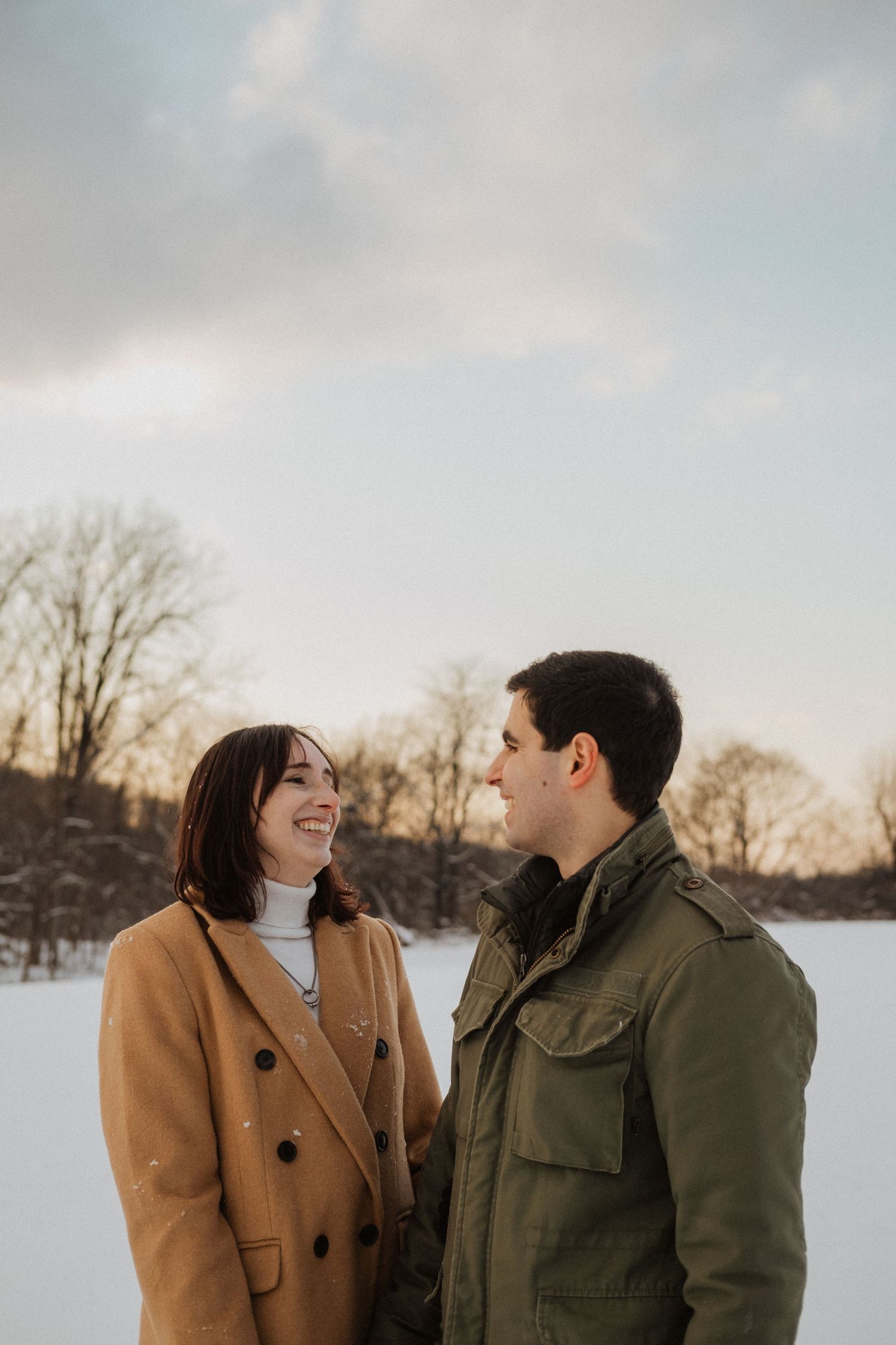 Smiling couple facing each other in a snowy winter landscape at dusk.