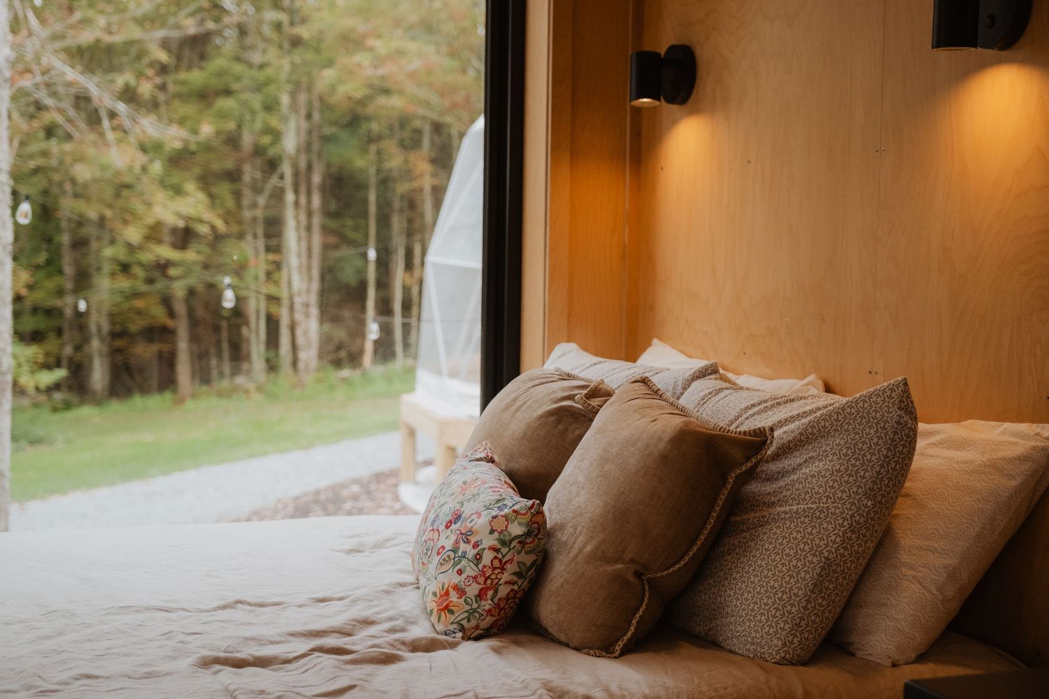 Cozy cabin bedroom with neutral pillows on a bed, wood-paneled walls, and large windows overlooking a lush green forest.