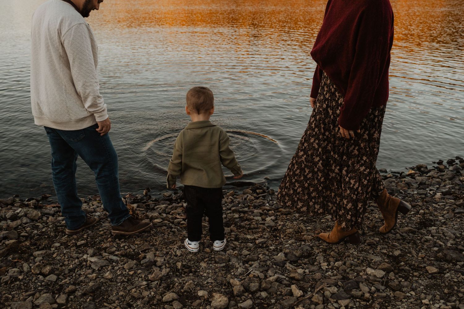 Family of three standing by a rocky lakeside at golden hour, toddler tossing a stone into calm water.
