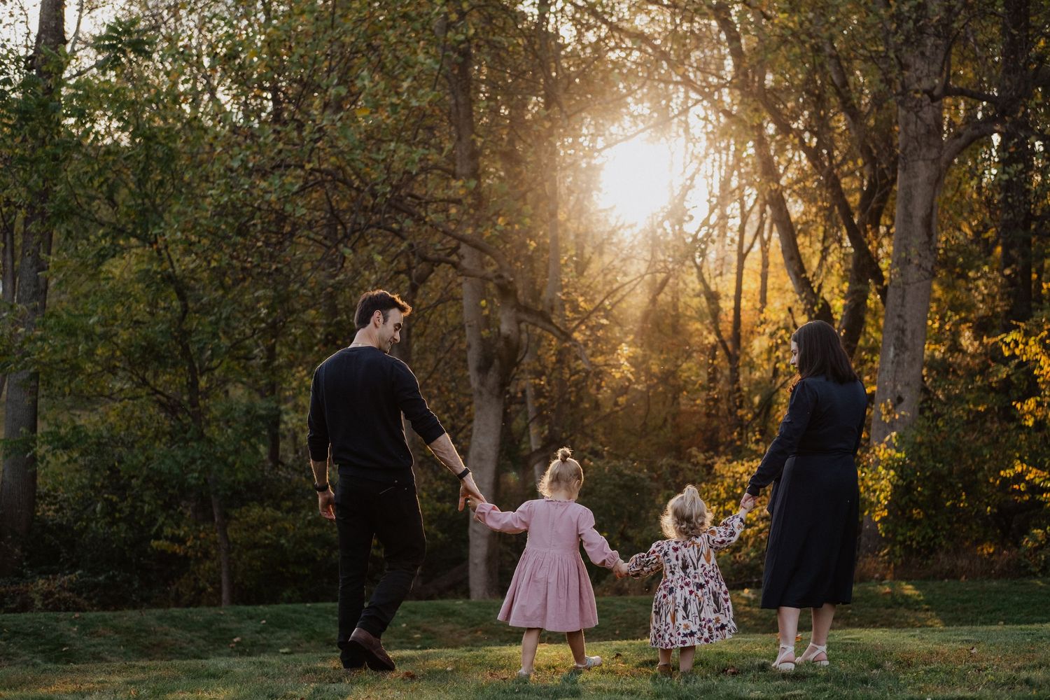 Family of four holding hands walking toward golden sunset light through autumn trees in a lush green park.