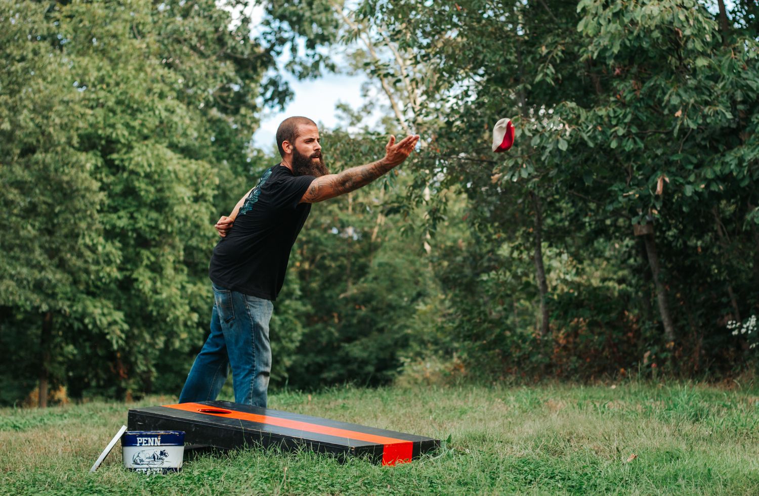 Man playing cornhole outdoors, tossing a red bean bag toward a black and orange board on a grassy lawn.