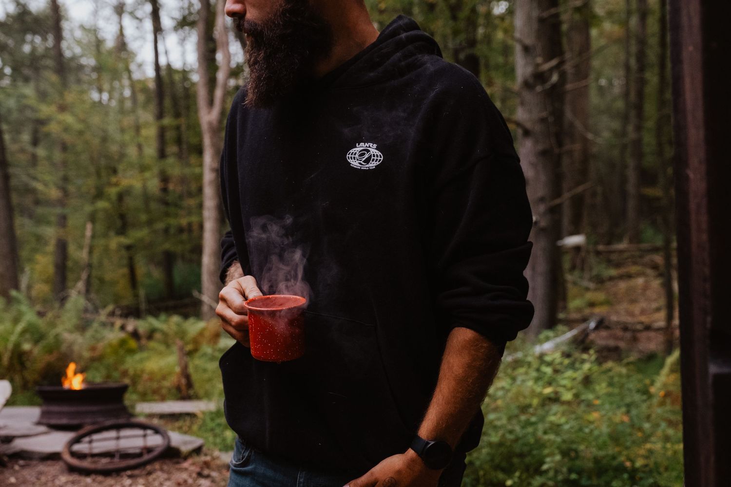 Man in black hoodie holding a steaming red mug outdoors in a forest campsite setting.