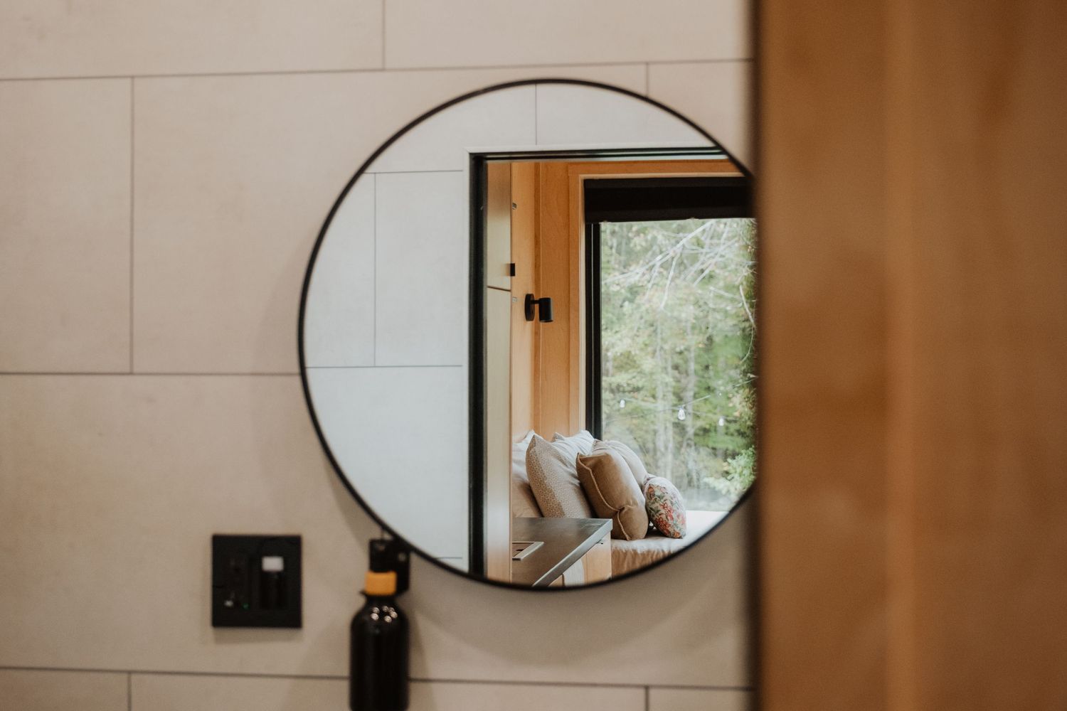 Round black-framed mirror reflecting a cozy window seat with cushions and lush green trees in a modern bathroom.