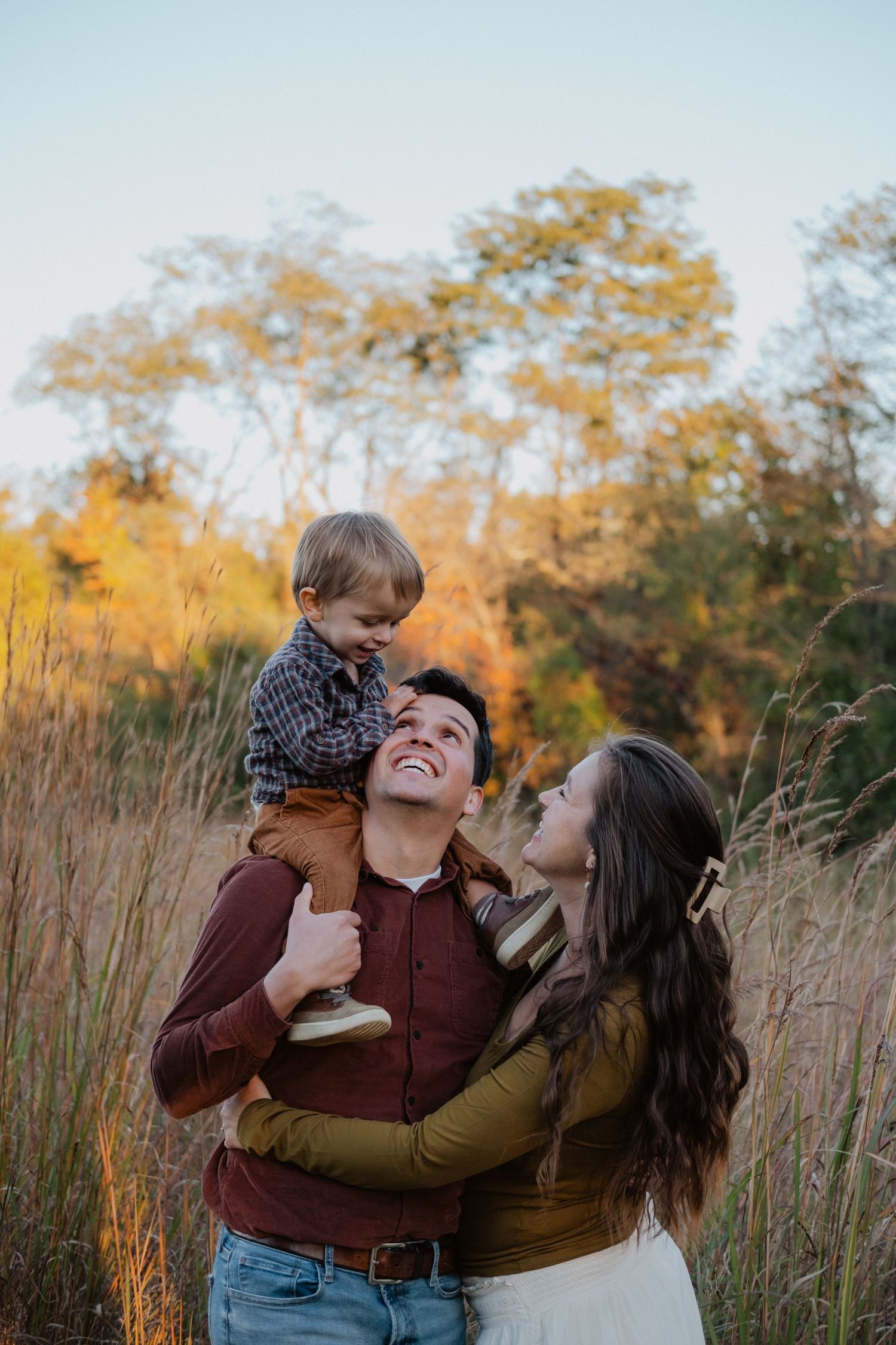 Happy family of three in autumn field, father carrying son on shoulders while mother embraces them both.