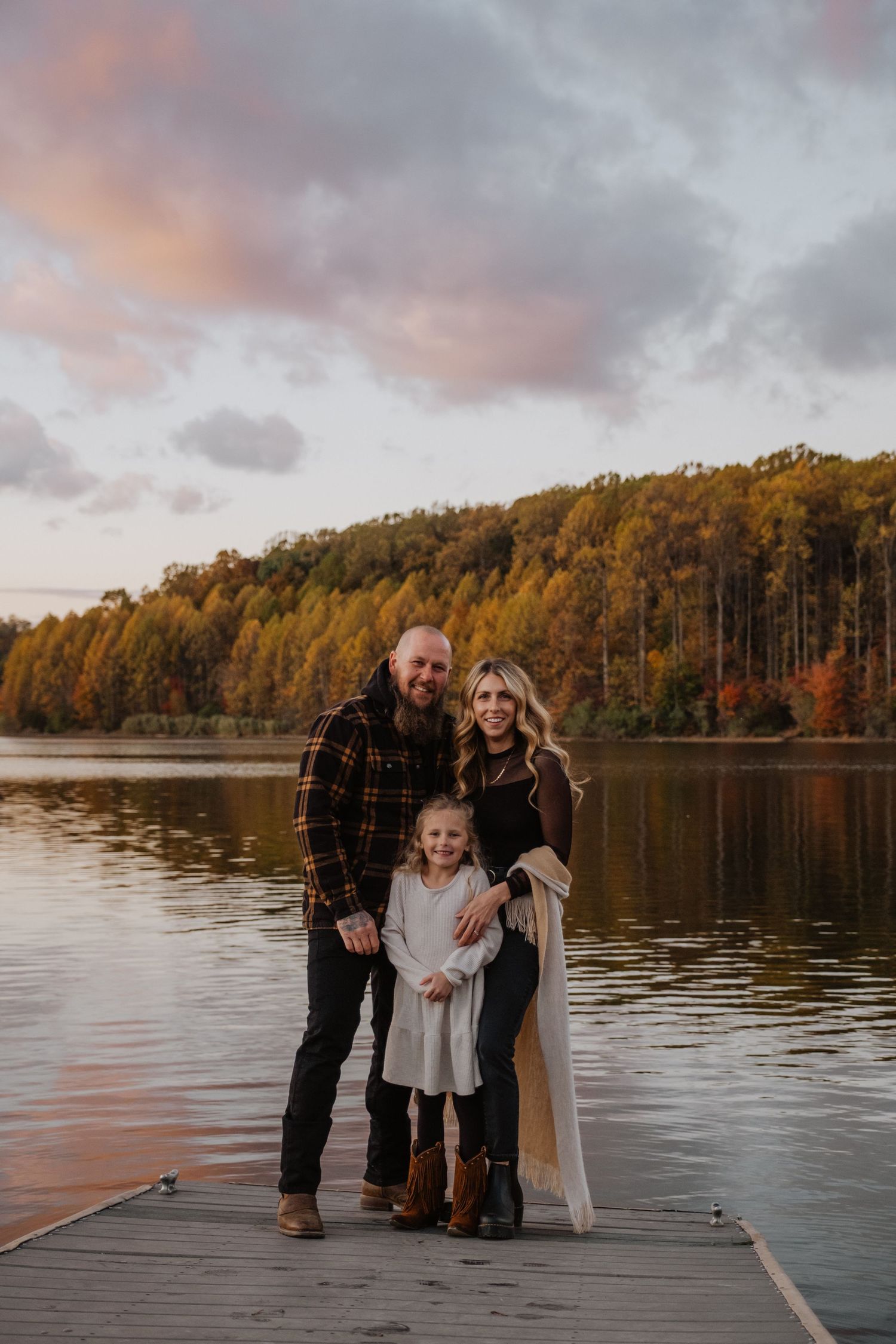 Family of three posing on a lake dock during golden hour with fall foliage in background.