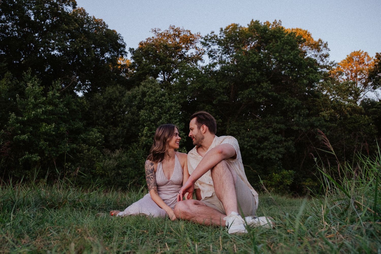 Romantic couple sitting together in a grassy field at dusk, gazing lovingly into each other's eyes.