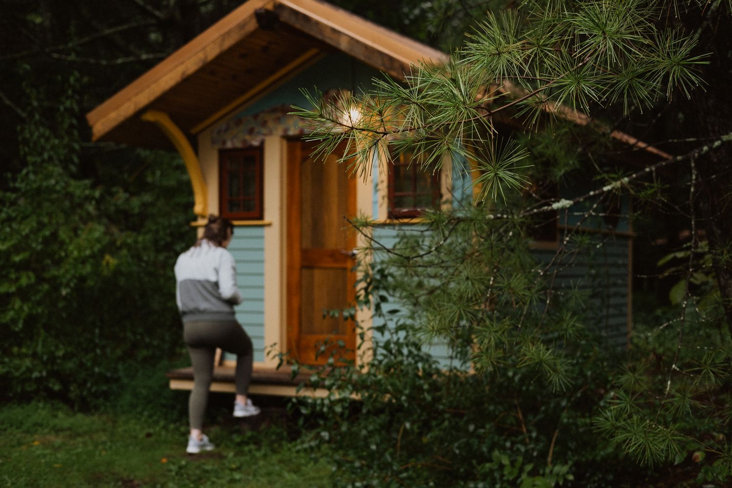 Woman sitting on bench outside a charming small wooden cabin surrounded by lush green trees and string lights.