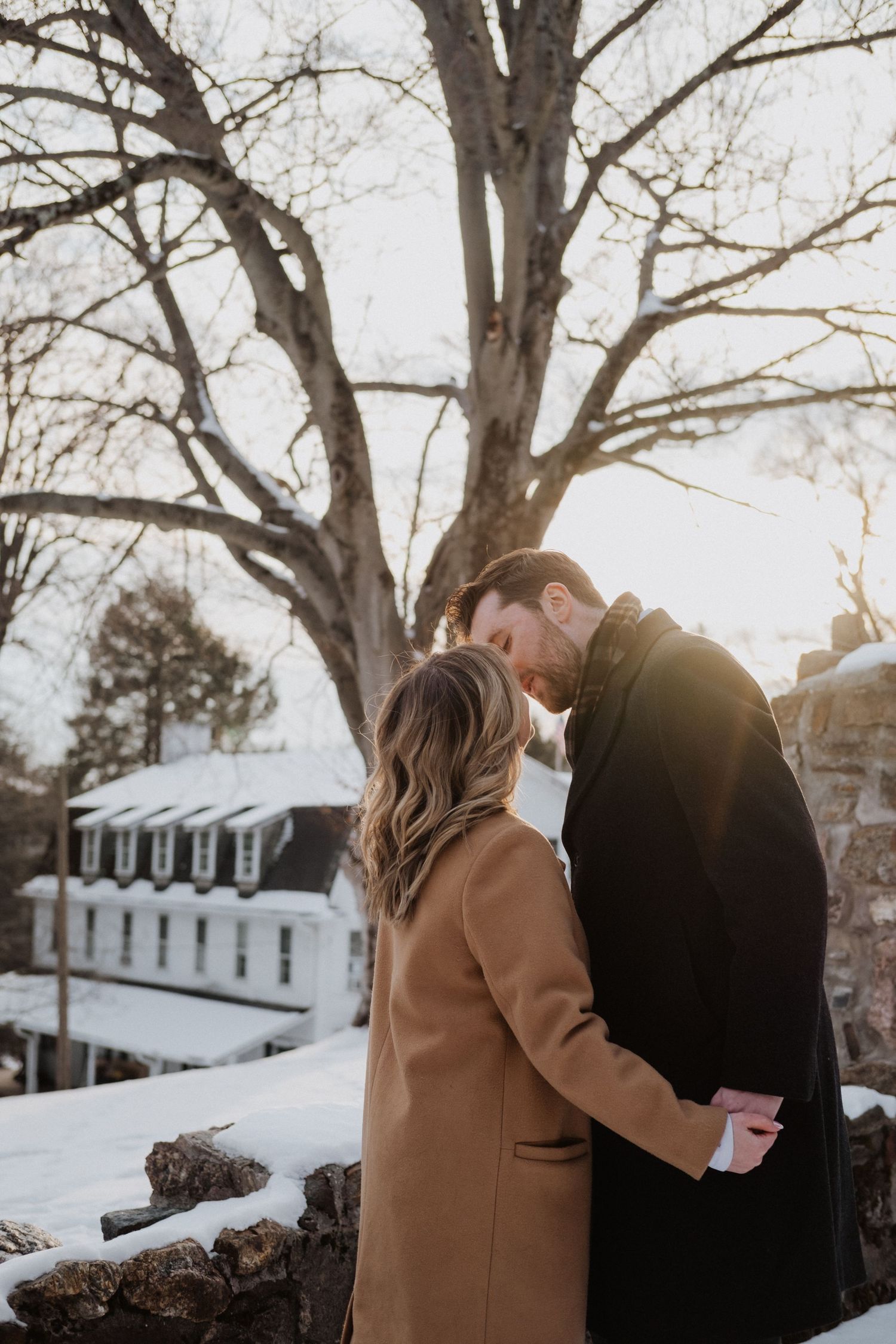 Couple sharing a romantic winter moment outdoors, dressed in elegant coats, with snow-covered ground and bare trees at sunset.
