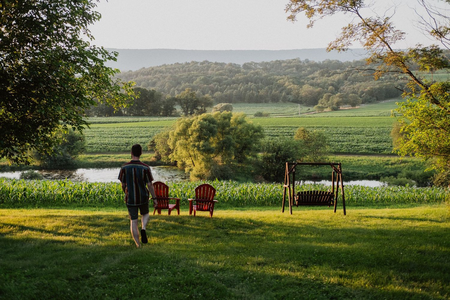 Person walking toward red chairs overlooking a lush green river valley with rolling hills at golden hour.