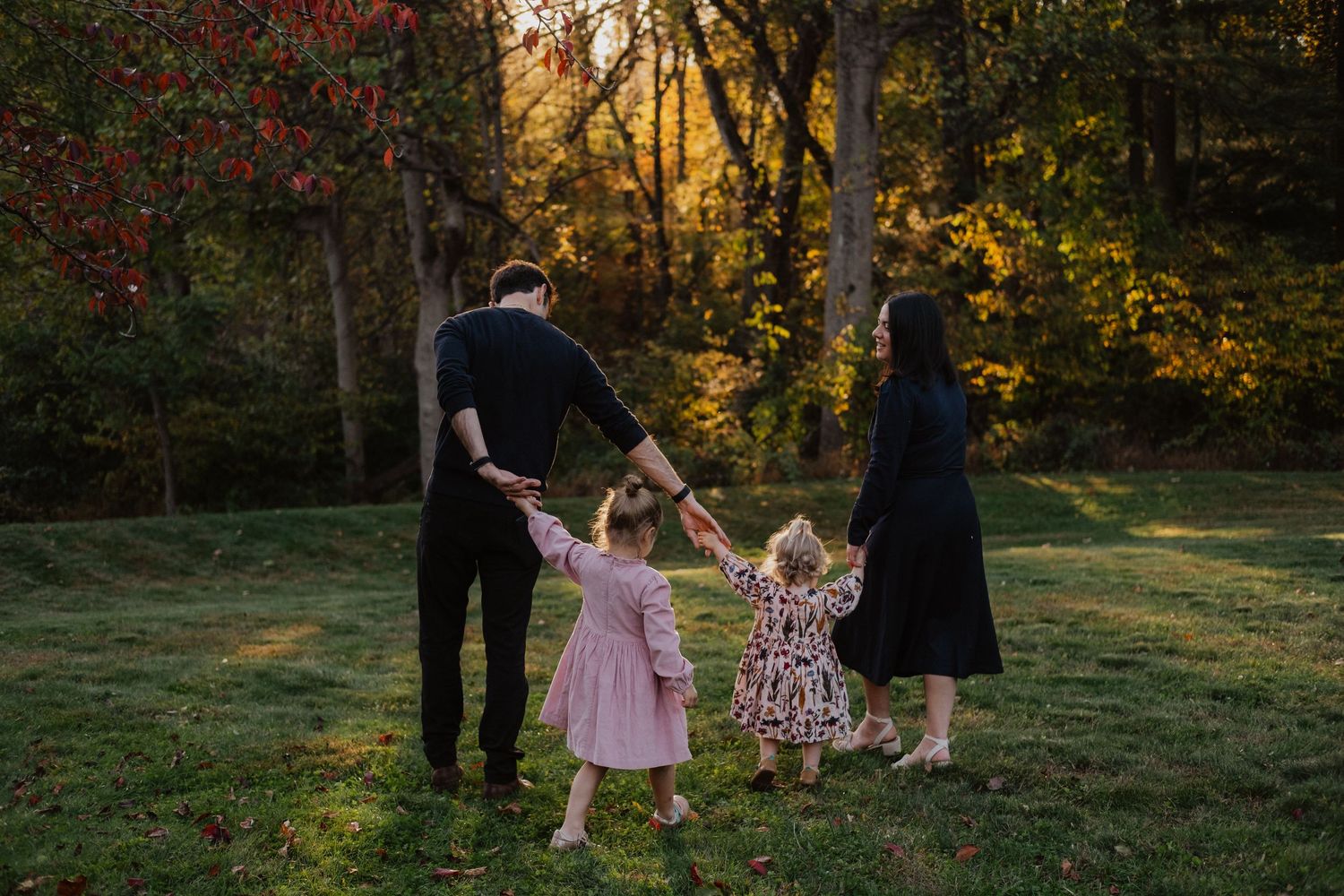 Family of four holding hands in circle among colorful autumn trees at golden hour.