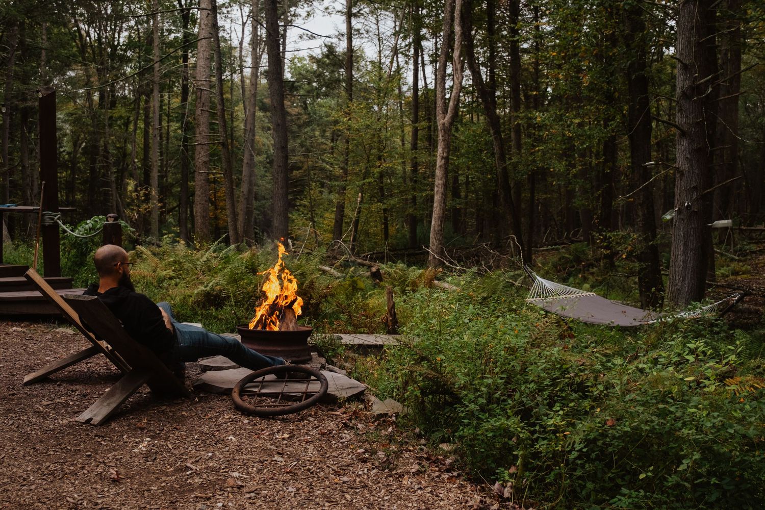 Person relaxing in a chair by a crackling campfire surrounded by lush green forest trees in a serene woodland setting.