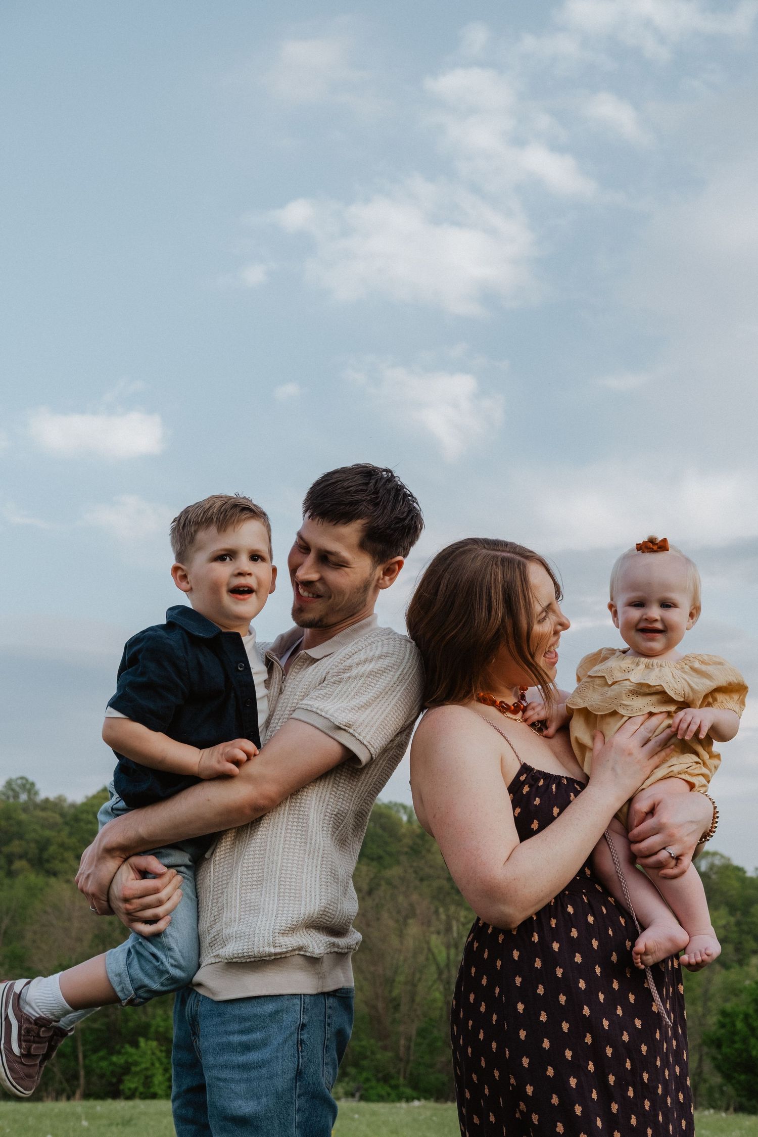 A happy family of four poses outdoors, parents holding two young children, smiling together against a blue sky.