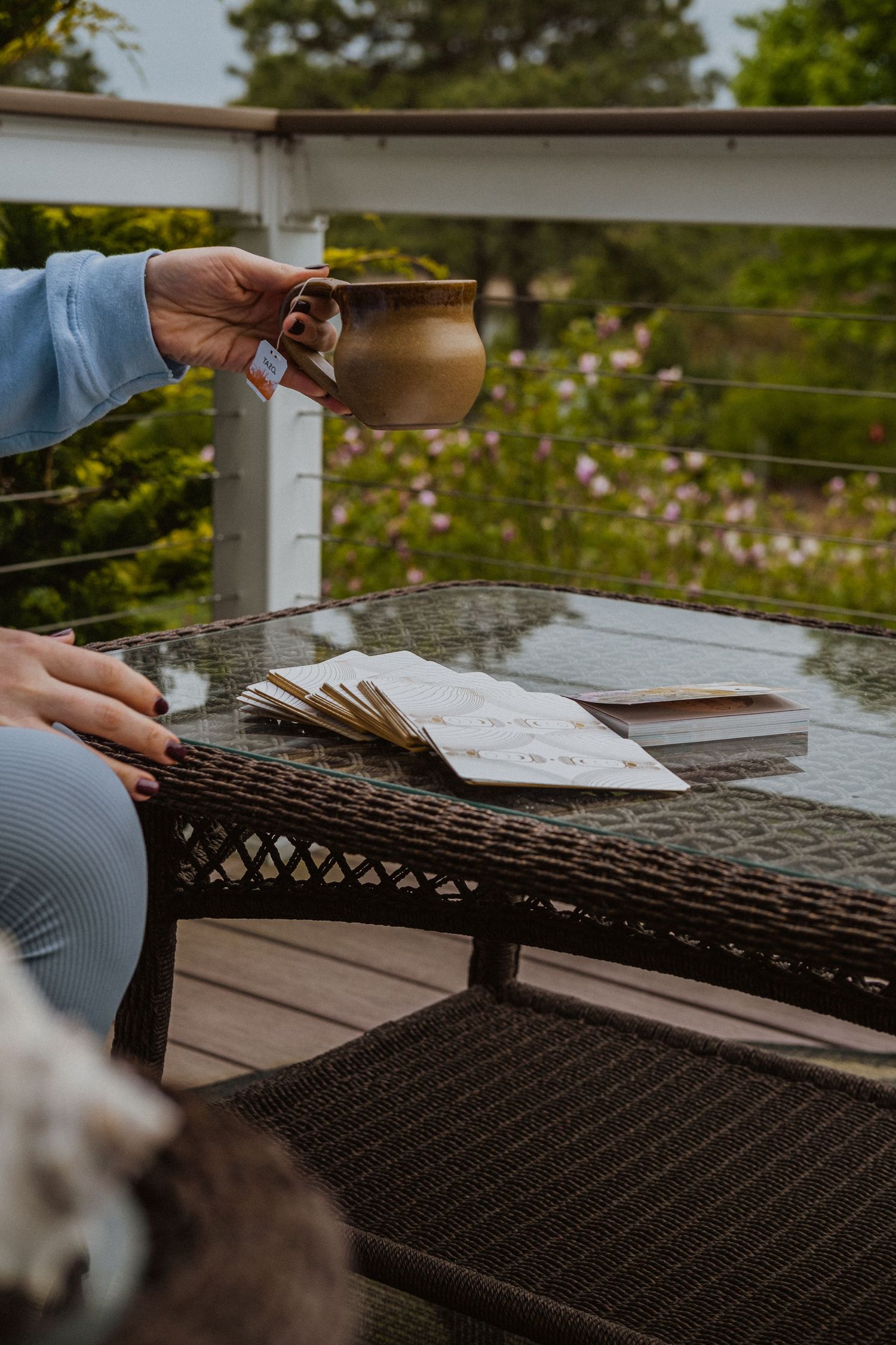 Woman holding a ceramic mug on an outdoor patio with wicker table, open books, and lush garden backdrop.
