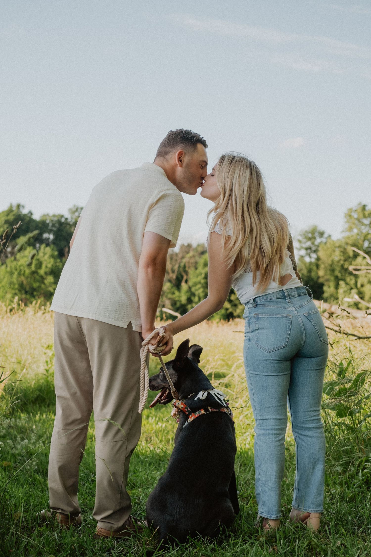 Couple sharing a kiss outdoors while their black dog sits between them.
