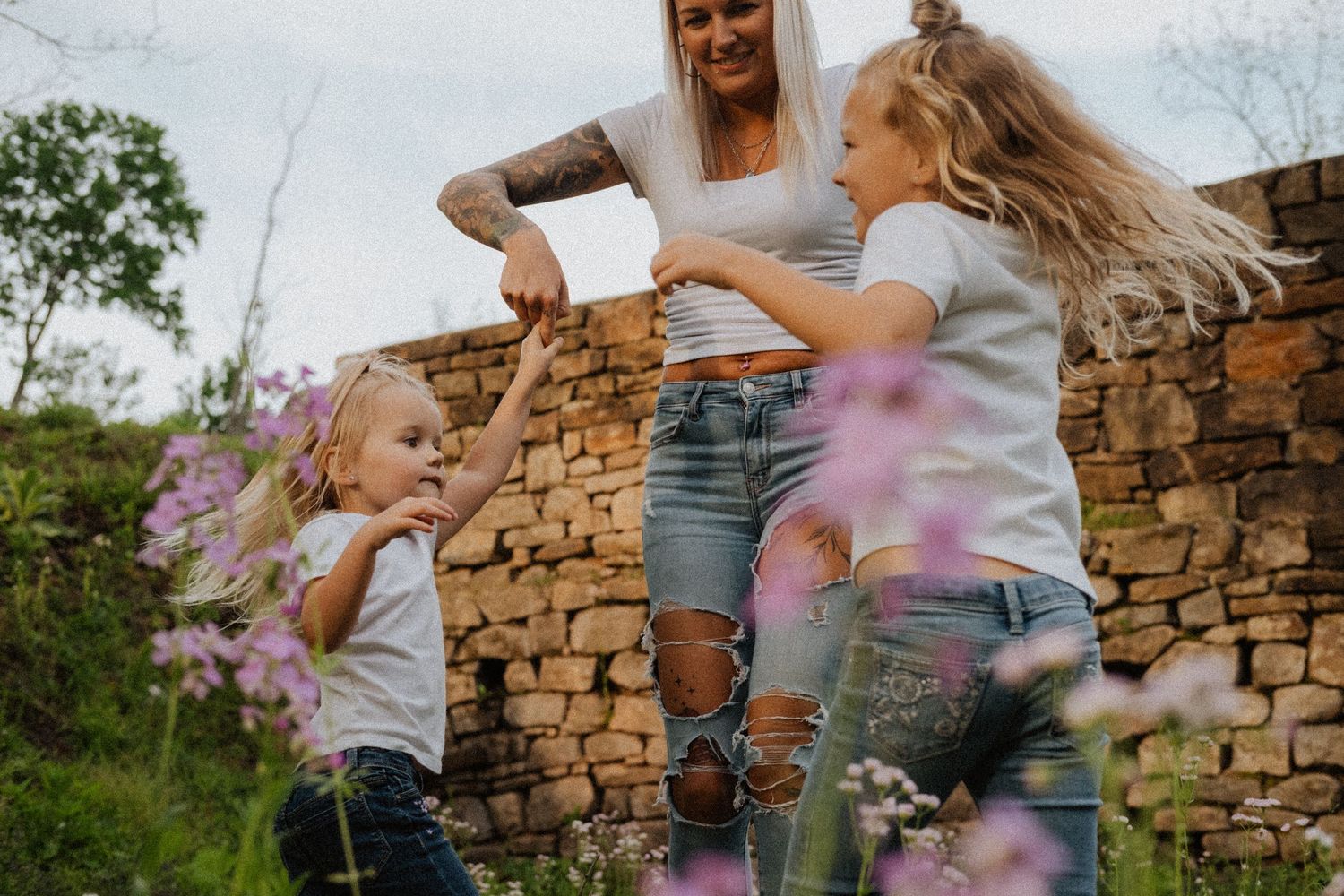 Tattooed mother playing and dancing with two young blonde girls near a rustic stone wall outdoors.