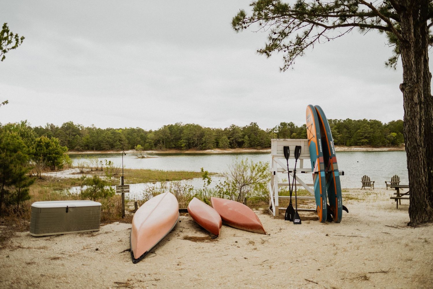 Kayaks and paddleboards rest on a sandy lakeside beach surrounded by lush greenery under an overcast sky.