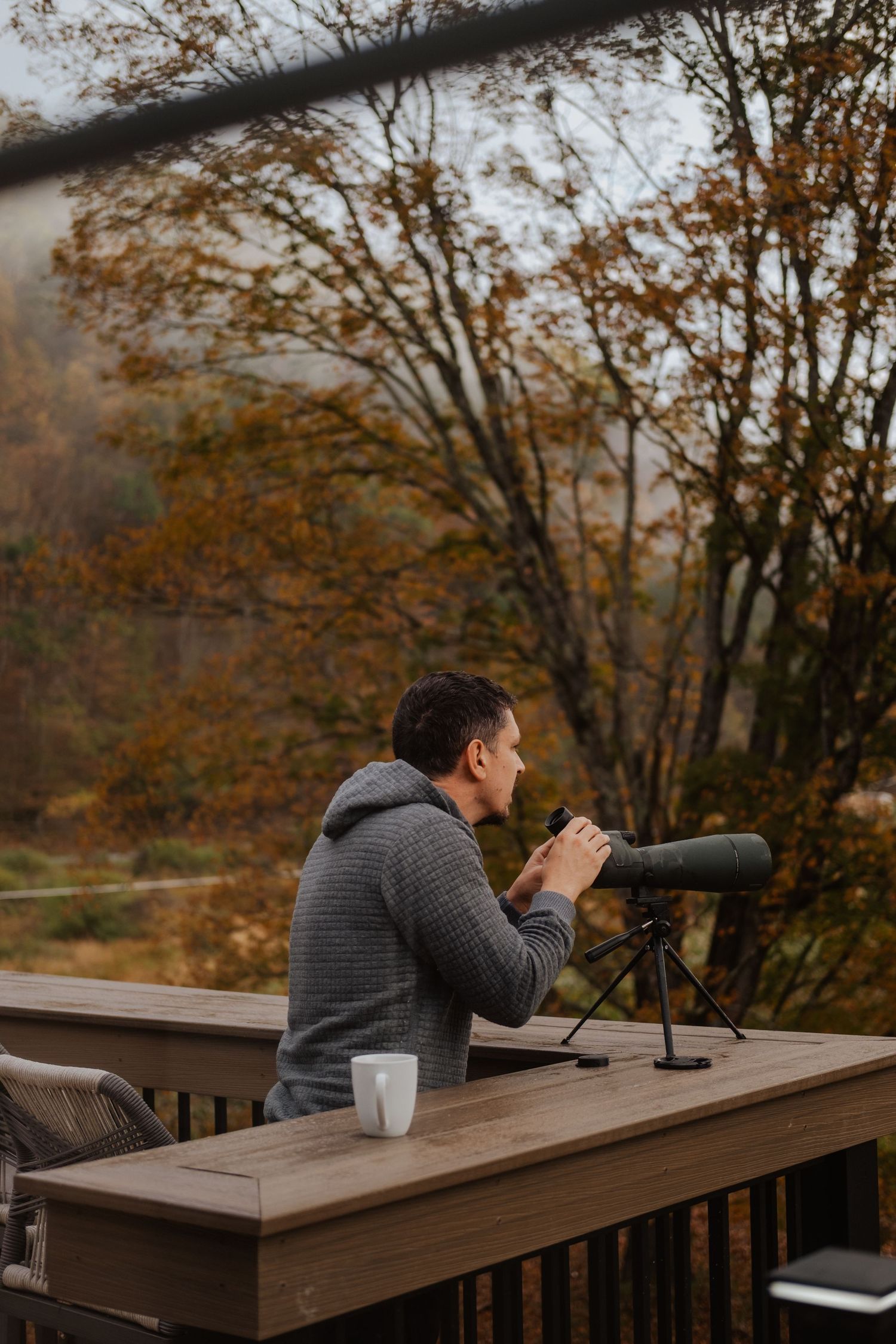 Man sitting on autumn deck with spotting scope and coffee mug, enjoying fall foliage views.