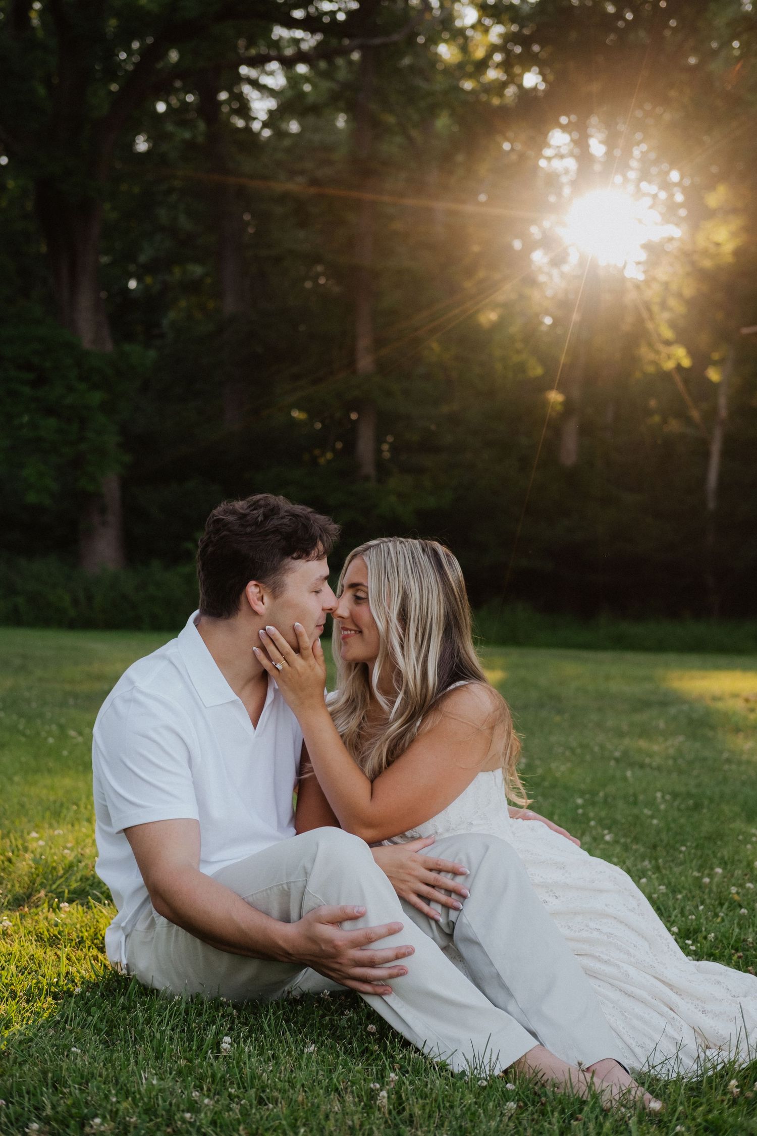 Couple in white outfits sitting on grass sharing a romantic moment at golden hour with sunlight filtering through trees.
