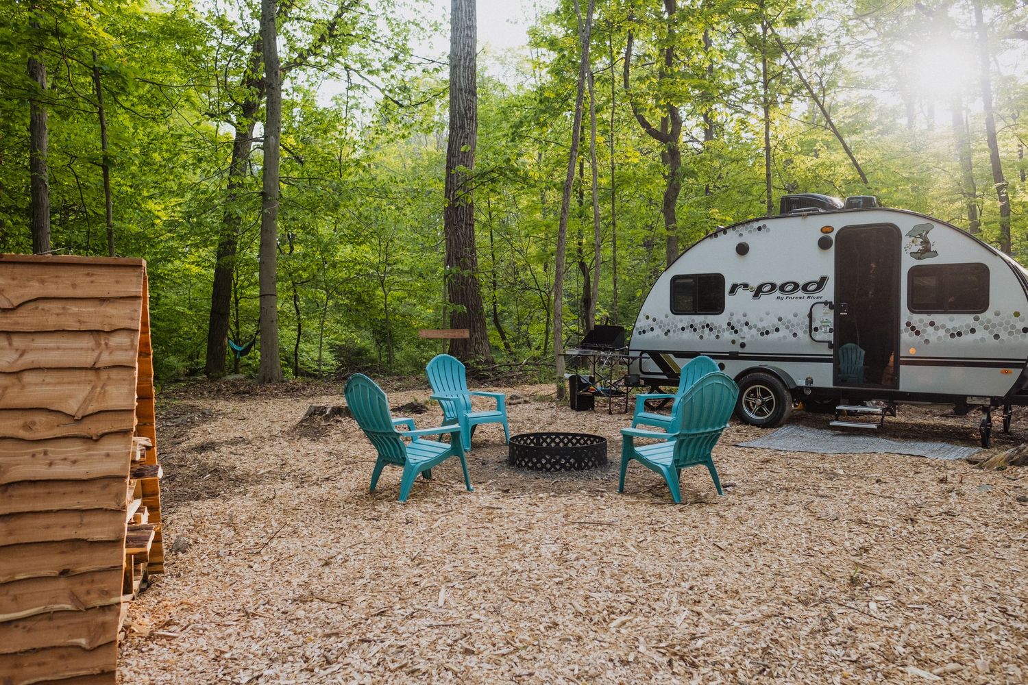 Cozy campsite with blue Adirondack chairs around a fire pit beside an r-pod trailer in a lush wooded forest.