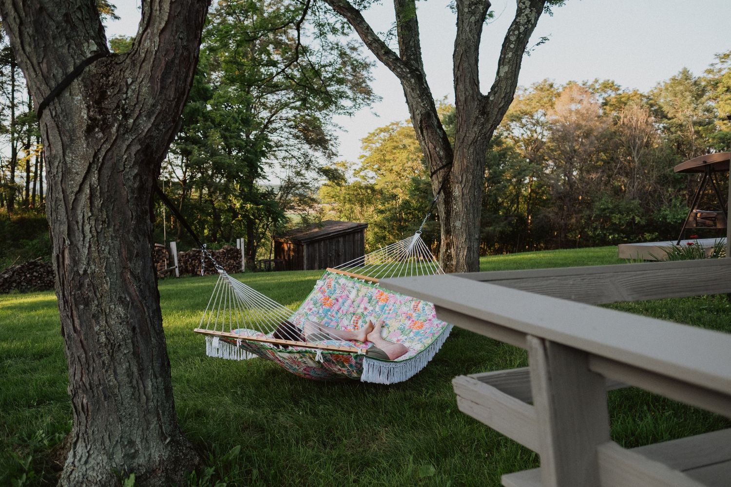A colorful hammock strung between two trees in a lush green backyard beside a wooden picnic table at dusk.