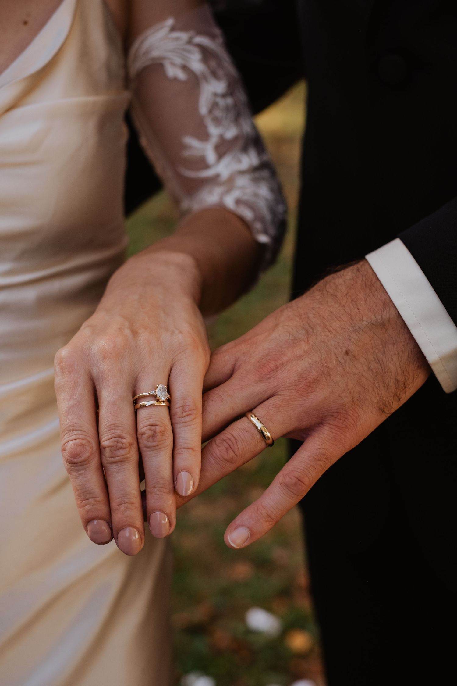 Close-up of newlyweds showing off wedding rings, bride in lace gown holding hands with groom in black suit.