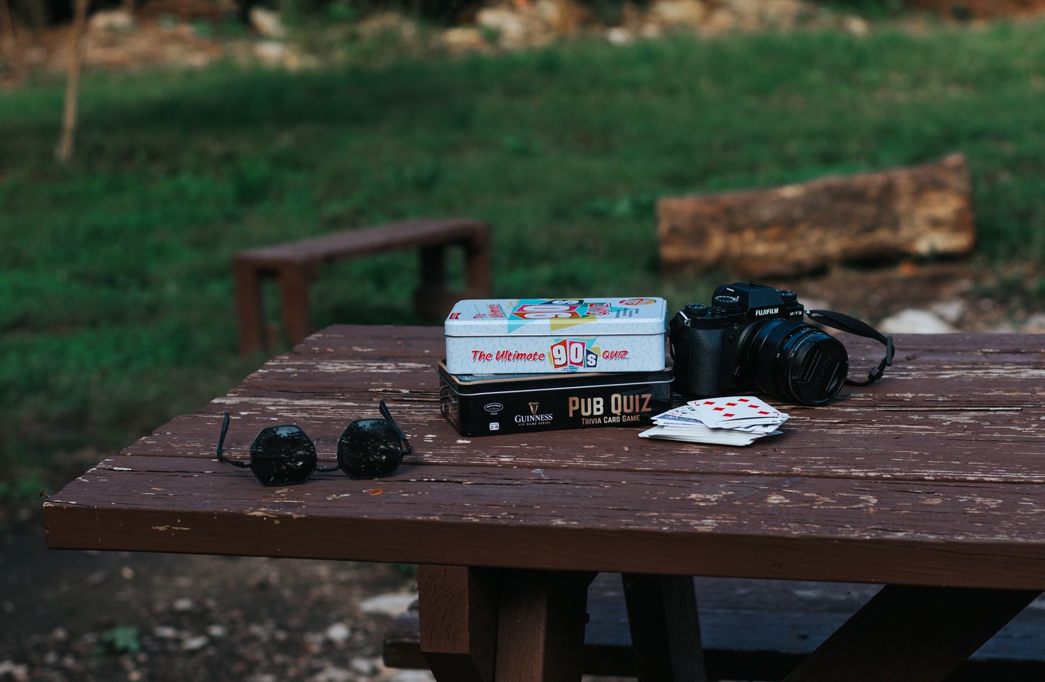 A camera, card game, and accessories arranged on a wooden picnic table outdoors with green grass in the background.