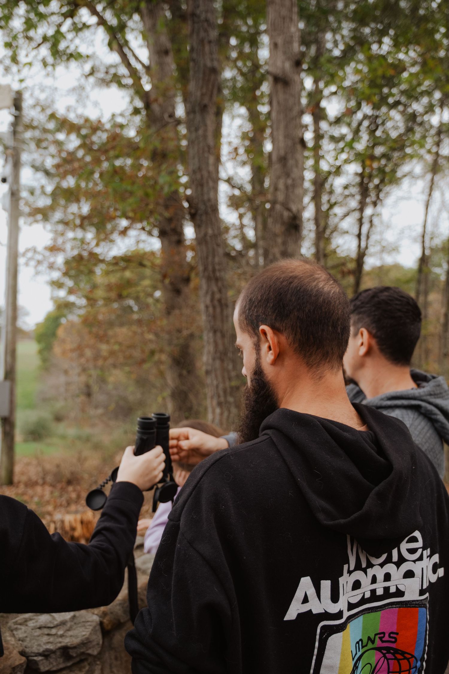 Men in hoodies stand outdoors near trees, one holding binoculars, wearing an Automatic Mars graphic sweatshirt.