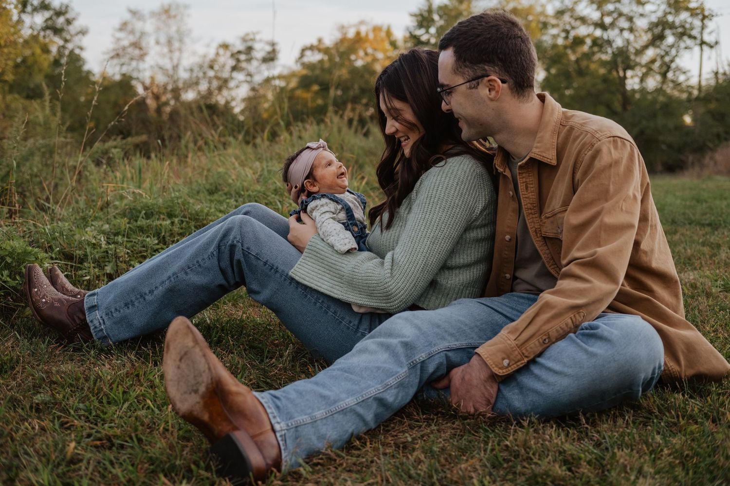 A couple sits on grass cradling their newborn baby outdoors in a lush green field during golden hour.