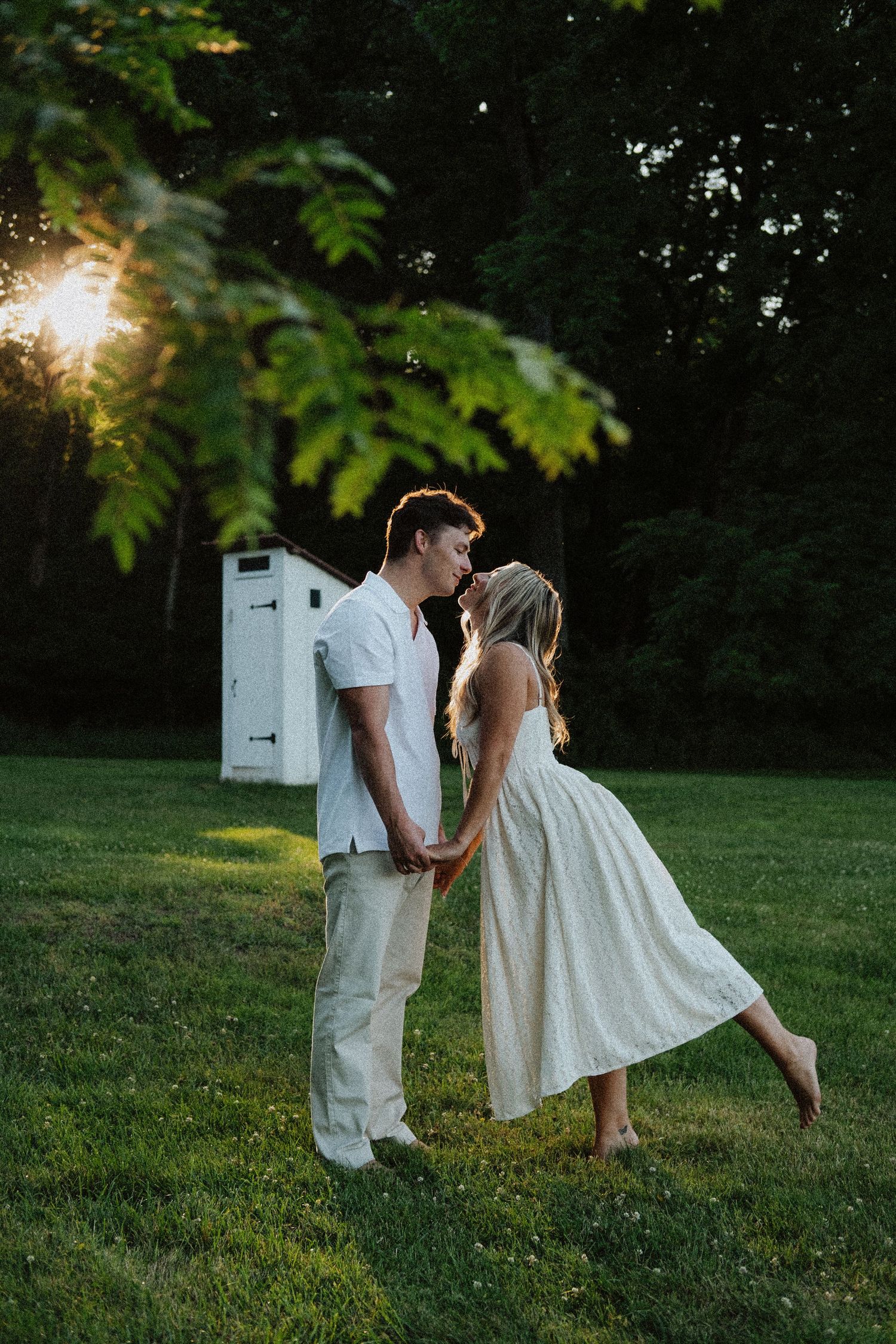 Couple sharing a tender kiss at sunset in a lush green garden, woman in white midi dress holding hands.