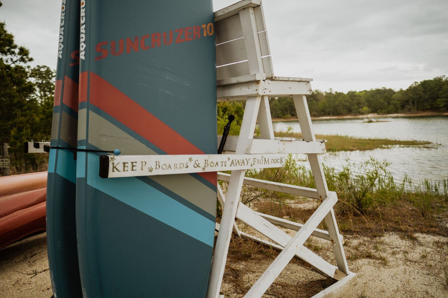 A white lifeguard chair stands beside a teal paddleboard on a sandy lakeside beach surrounded by trees.