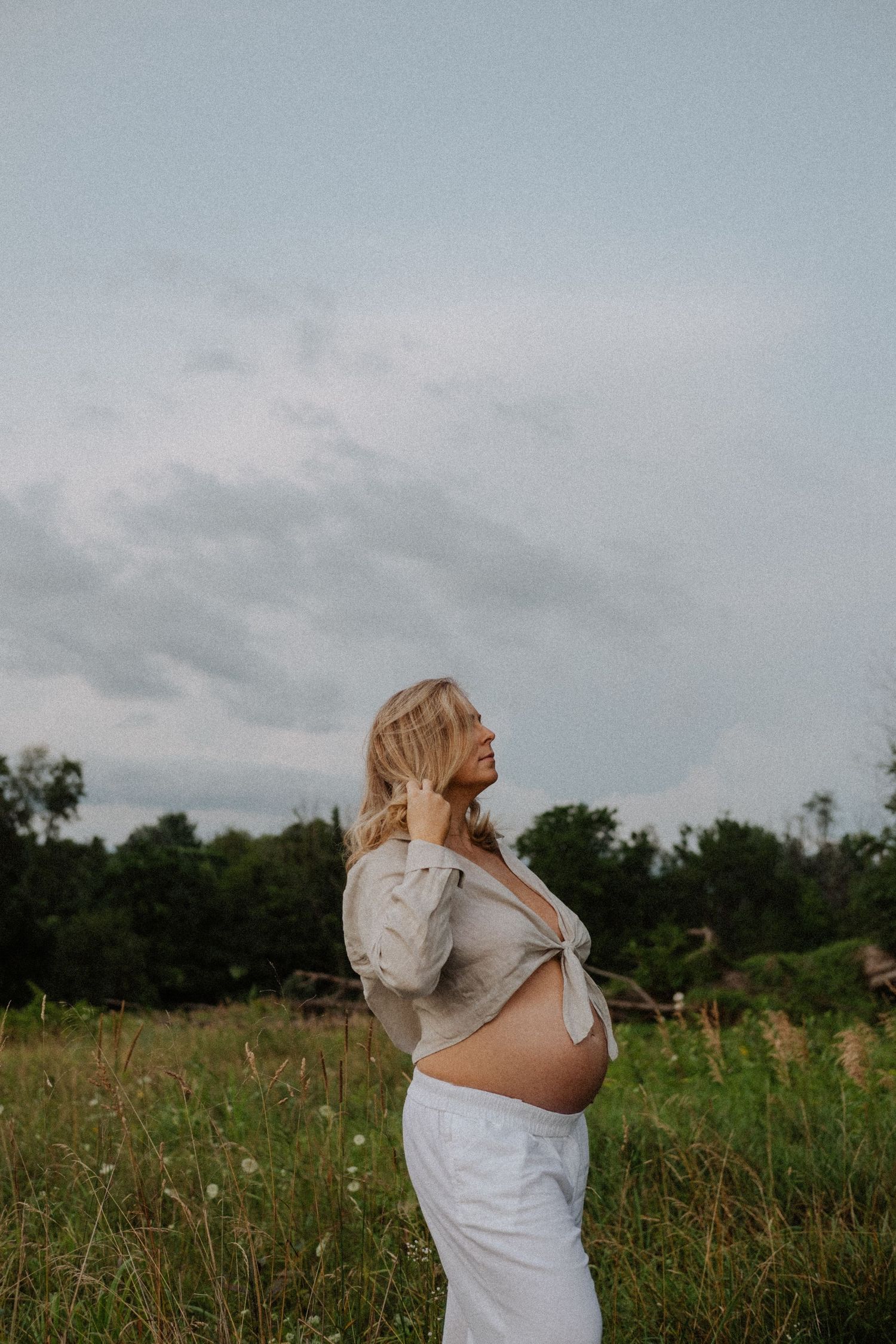 Pregnant woman in white pants and tied beige top stands in a green field, showing baby bump under cloudy sky.