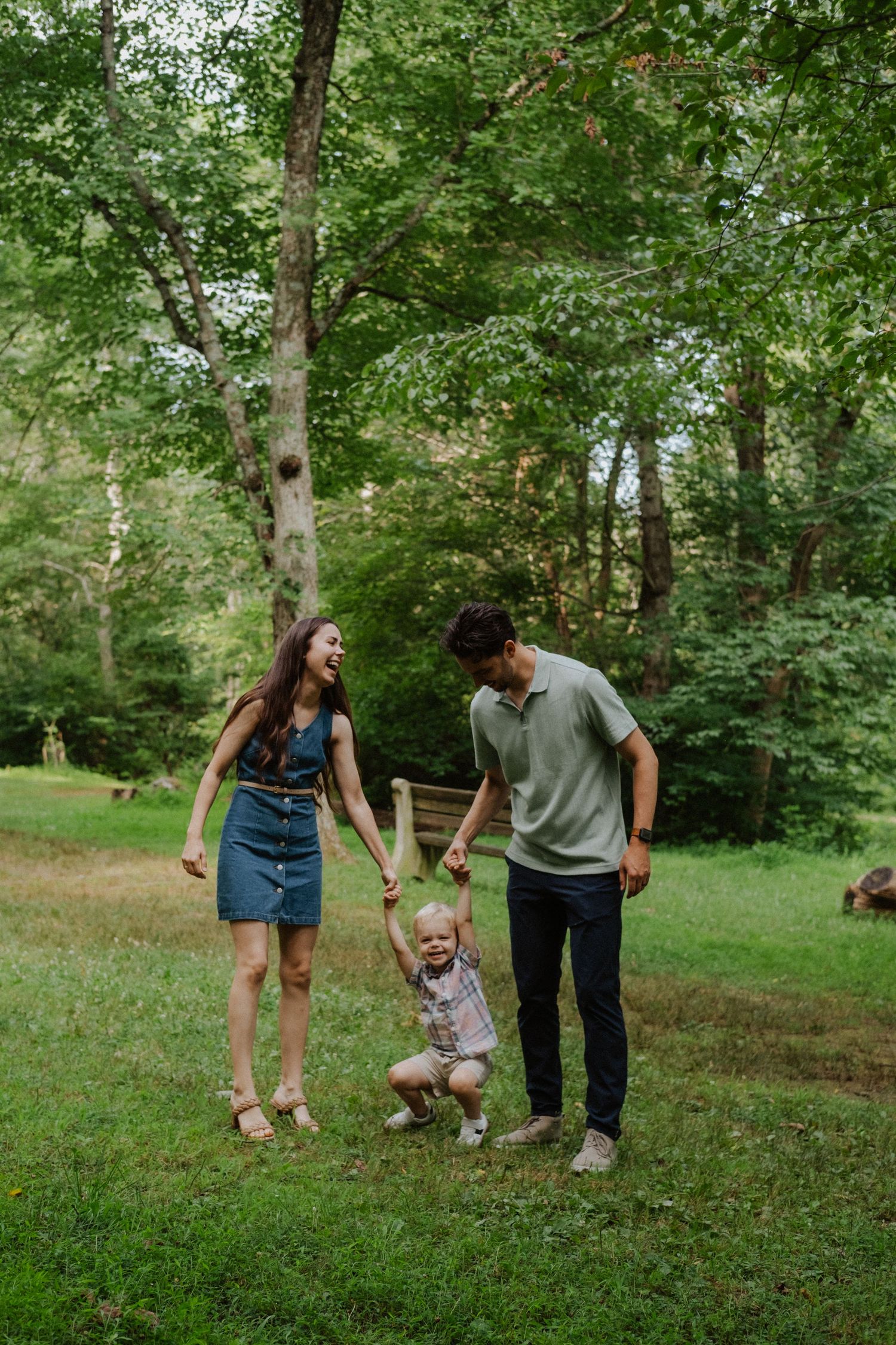 Young family playing together in a forest clearing, parents swinging toddler by hands on green grass.