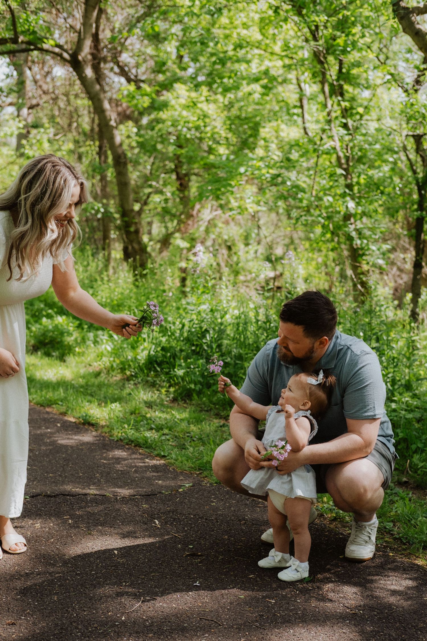 Father crouching with baby girl as mother offers wildflowers on lush green forest path.